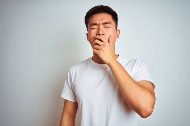 İzole edilmiş beyaz arka plan üzerinde duran Asyalı genç adam esnemekten sıkılmış, elleriyle ağzını kapatmış. Huzursuz ve uykulu..