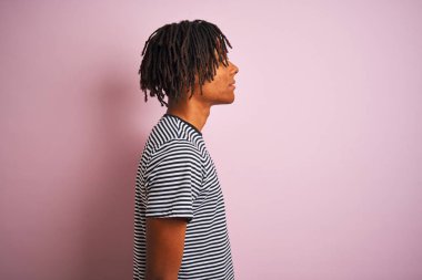 Afro man with dreadlocks wearing navy striped t-shirt standing over isolated pink background looking to side, relax profile pose with natural face with confident smile.