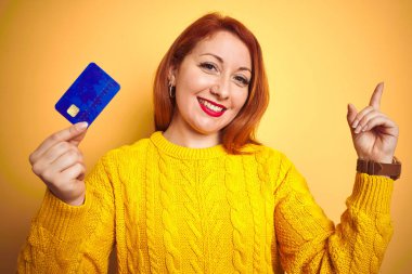 Genç müşteri kızıl saçlı kadın kredi kartını sarı izole edilmiş arka planda tutarken mutlu bir şekilde elini ve parmağını kenarı işaret ediyor.