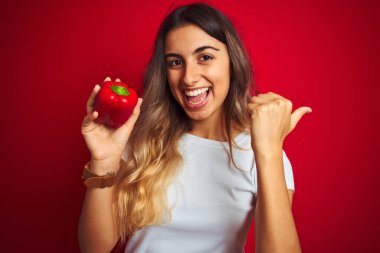 Kırmızı izole edilmiş arka planda biber tutan genç ve güzel bir kadın başparmağıyla gülümseyerek yüzünü gösteriyor.