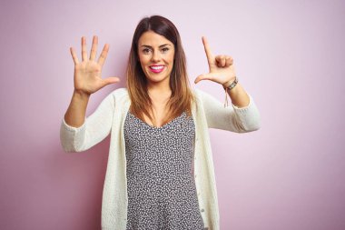 Pembe izole edilmiş arka planda duran genç ve güzel bir kadın yedinci parmaklarını gösterip işaret ederken kendinden emin ve mutlu gülümsüyor..