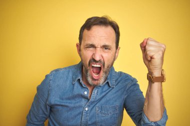 Yakışıklı, orta yaşlı, gri saçlı, izole edilmiş sarı arka planı olan öfkeli ve öfkeli sinirli bir adam öfkeyle bağırırken yumruğunu kaldırıyor. Öfke ve agresif kavram.
