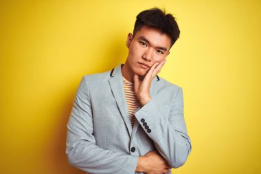 Genç Asyalı Çinli iş adamı, sarı arka plan üzerinde duran ceketini giyer ve kollarını kavuşturarak depresyondan sıkılmış ve yorgun gözükür..