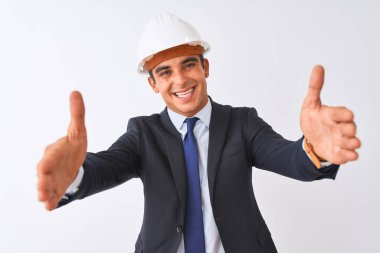 Takım elbiseli ve kasklı genç bir mimar izole edilmiş beyaz arka planda kameraya bakarak sarılmak için kollarını açmış gülümsüyor. Mutluluğu kucaklayan neşeli bir ifade..