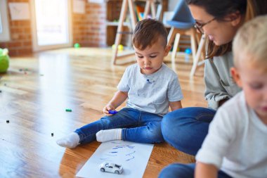 Anaokulunda bir sürü oyuncağın etrafında kağıt ve kalem kullanarak yerde oturan bir grup güzel öğretmen ve çocuk.