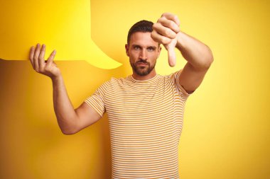 Genç adam, sarı izole edilmiş arka planda mesaj için boş konuşma balonu tutuyor. Kızgın yüzlü, olumsuz işaretli, başparmakları aşağıda, reddedilme kavramlı.