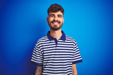 Yüzünde mutlu ve havalı bir gülümsemeyle izole edilmiş mavi arka planda duran çizgili polo giyen genç bir adam. Şanslı kişi..