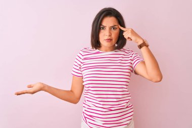 İzole edilmiş pembe arka planda duran çizgili tişört giyen genç ve güzel bir kadın. Kafası karışmış ve avuç içi açık bir şekilde fotokopi alanı ve alnını işaret etmekten rahatsız olmuş. Bunu bir düşün..