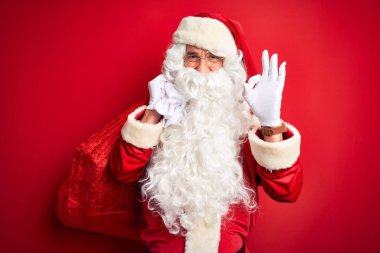 Orta yaşlı bir adam Noel Baba kostümü giyiyor. Kırmızı arka planda hediyeler taşıyor. Tamam işaretini parmaklarıyla yapıyor.