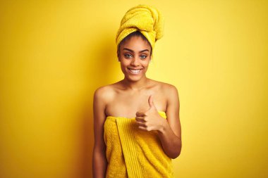 Genç Afrikalı Amerikalı kadın duş üstüne havlu giyip izole edilmiş sarı arka planda mutlu bir şekilde gülümsüyor.