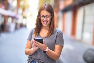 Mutlu ve kendinden emin gülümseyen genç ve güzel bir kadın. Yüzünde gülümsemeyle şehrin caddesinde akıllı telefon kullanarak dikiliyorsun.