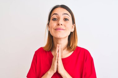 Güzel kızıl saçlı kadın, rahat kırmızı tişörtler giyip izole edilmiş arka planda dua ediyor ellerinin birliğiyle af diliyor kendinden emin bir şekilde gülümsüyor..