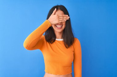 Turuncu tişört giyen genç, güzel Çinli bir kadın izole edilmiş mavi arka planda duruyor. Gülümsüyor ve elleri yüzünde şaşkınlıktan gözleri kapatıyordu. Kör kavram.