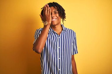 Rastgele çizgili tişört giyen Afro bir adam izole edilmiş sarı arka planda bir gözünü kapatıyor, yüzünde kendine güvenen bir gülümseme ve şaşırtıcı bir duygu var..