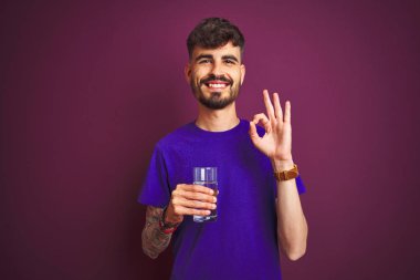 Dövmeli genç adam izole edilmiş mor arka planda duran bir bardak su içiyor. Tamam işareti yapıyor, mükemmel bir sembol.