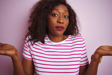 Çizgili tişört giyen Afro-Amerikalı genç bir kadın soyutlanmış pembe arka plan üzerinde duruyor habersiz ve elleri ve kolları havada kafası karışmış bir ifade. Şüphe kavramı.
