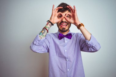 Mor gömlekli ve papyonlu genç adam izole edilmiş beyaz arka planda dürbün gibi güzel hareketler yapıyor. Dürbün dilini dışarı çıkarıyor, gözler parmaklarından bakıyor. Çılgın ifade.