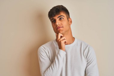 İzole edilmiş beyaz arka plan üzerinde duran kazak giyen genç Hintli adam elleri çenesinde soru sorarken düşünceli bir ifade takınıyor. Düşünceli bir yüzle gülümsüyor. Şüphe kavramı.