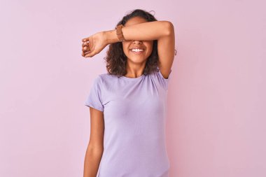 İzole edilmiş pembe arka planda duran tişört giyen genç Brezilyalı bir kadın. Gözleri kollarıyla kapatıp neşeli ve komik bir şekilde gülümsüyor. Kör kavram.