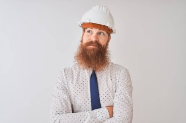 Genç kızıl saçlı mimar, izole edilmiş beyaz arka planda güvenlik kaskı takıyor. Gülümsüyor ve başka tarafa bakıyor..