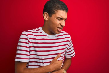 Genç yakışıklı Arap adam izole edilmiş kırmızı arka plan üzerine çizgili tişört giyiyor. Midesinde hazımsızlık, acı verici bir hastalık var. Ache kavramı.