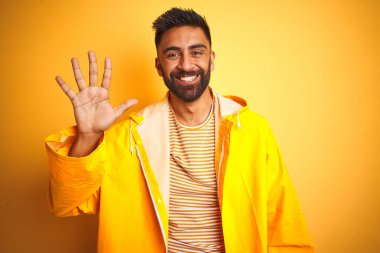 İzole edilmiş sarı arka planda duran yağmurluk giyen genç Hintli adam beş numaralı parmaklarıyla işaret ederken kendinden emin ve mutlu bir şekilde gülümsüyor..