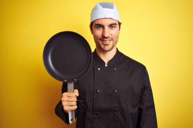 Üniformalı ve şapkalı bir aşçı şapkalı genç adam izole edilmiş sarı arka planda mutlu bir yüzle ayakta duruyor ve gülümsüyor dişlerini gösteren bir gülümsemeyle.