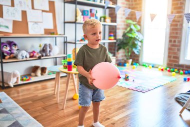 Anaokulunda pembe balonla oynayan beyaz bir çocuk. Anaokulu çocuğu oyuncaklarla oyun odasında mutlu.