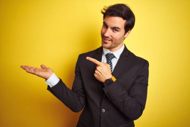 Takım elbiseli ve kravatlı genç yakışıklı işadamı, izole edilmiş sarı arka planda durup kameraya hayret ediyor ve gülümsüyor..