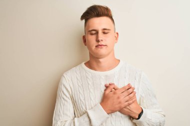 Kışlık süveter giyen genç bir adam soyutlanmış beyaz arka plan üzerinde duruyor elleri göğsünde, gözleri kapalı ve yüzünde minnet dolu bir jest ile. Sağlık kavramı.