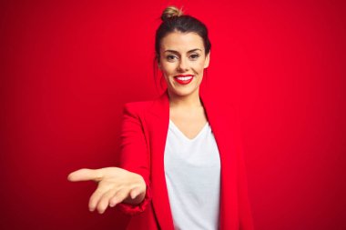 Young beautiful business woman standing over red isolated background smiling cheerful offering palm hand giving assistance and acceptance. clipart
