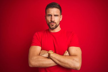 Genç yakışıklı adam kırmızı izole edilmiş arka plan şüphecisi ve gergin kolları çaprazlanmış yüz ifadesini tasvip etmeyen sıradan bir tişört giyiyor. Negatif kişi.