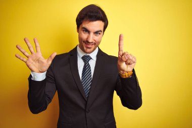 Takım elbiseli ve kravatlı genç yakışıklı işadamı izole edilmiş sarı arka planda duruyor ve altı numaralı parmaklarıyla işaret ederken kendinden emin ve mutlu gülümsüyor..