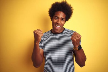 Afro saçlı, donanma çizgili tişörtlü, izole edilmiş sarı arka plan üzerinde afro-amerikalı bir adam kollarını kaldırarak ve açık gözlerle başarıyı kutluyor ve şaşırıyor. Kazanan konsept.