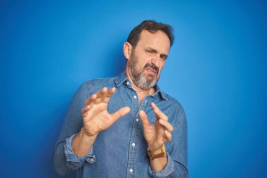 Yakışıklı, orta yaşlı, gri saçlı, soyutlanmış mavi arka planlı, tiksindirici, tiksindirici, tiksindirici bir yüz ifadesi olan yaşlı bir adam. Eller havada. Can sıkıcı bir kavram..