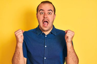 Kotlu tişört giyen genç adam, izole edilmiş sarı arka planda durup kollarını kaldırarak ve açık gözlerle başarının şaşkınlığını kutluyor. Kazanan konsept.