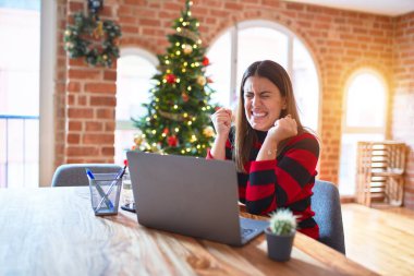 Güzel bir kadın evinde dizüstü bilgisayarla oturmuş Noel ağacının etrafında mutlu ve heyecanlı bir şekilde kolları havada, gülümseyerek ve başarı için çığlık atarak kazanan jestler yapıyor. Kutlama konsepti.