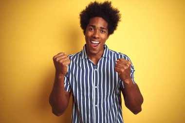 Afro saçlı, çizgili tişörtlü Amerikalı bir adam izole edilmiş sarı arka planda dikilmiş kollarını kaldırarak ve açık gözlerle başarıyı kutluyor ve şaşırıyor. Kazanan konsept.