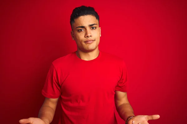 年轻的巴西男子穿着T恤 站在孤立的红色背景之上 毫无头绪 与张开的双臂混淆不清 没有概念 — 图库照片