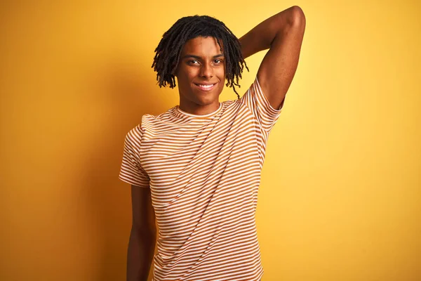 Afro man with dreadlocks wearing striped t-shirt standing over isolated yellow background smiling confident touching hair with hand up gesture, posing attractive and fashionable
