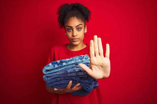 Young african american woman holding stack of jeans over isolated red background with open hand doing stop sign with serious and confident expression, defense gesture