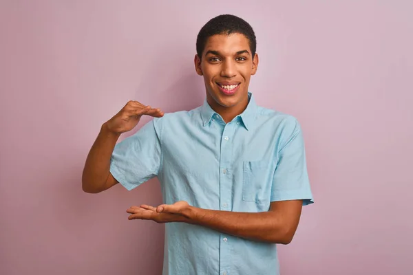 年轻英俊的阿拉伯男子 身穿蓝色衬衫 站在孤立的粉色背景上 手举着大号和大号的标志 象征着什么 笑着看着摄像机 计量概念 — 图库照片