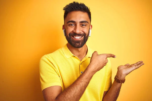 Young indian man listening to music using earphones standing over isolated yellow background amazed and smiling to the camera while presenting with hand and pointing with finger.