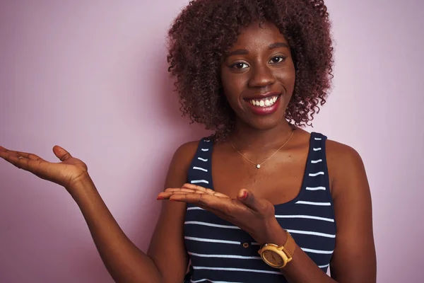 Young african afro woman wearing striped t-shirt standing over isolated pink background amazed and smiling to the camera while presenting with hand and pointing with finger.