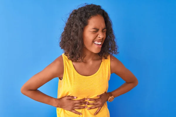 Young brazilian woman wearing yellow t-shirt standing over isolated blue background with hand on stomach because indigestion, painful illness feeling unwell. Ache concept.