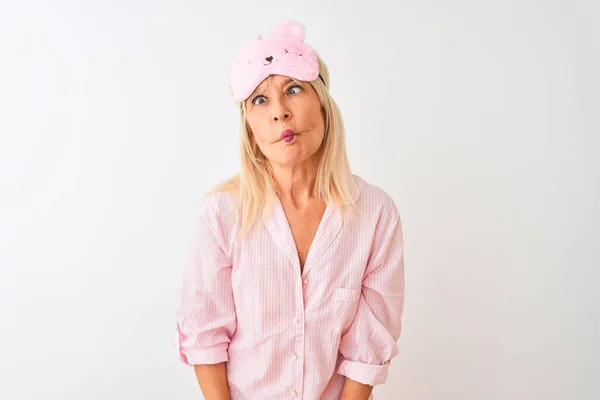 Middle age woman wearing sleep mask and pajama over isolated white background making fish face with lips, crazy and comical gesture. Funny expression.