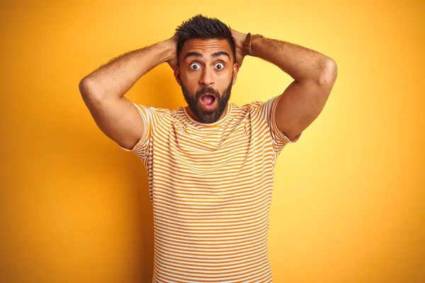 Young indian man wearing t-shirt standing over isolated yellow background Crazy and scared with hands on head, afraid and surprised of shock with open mouth