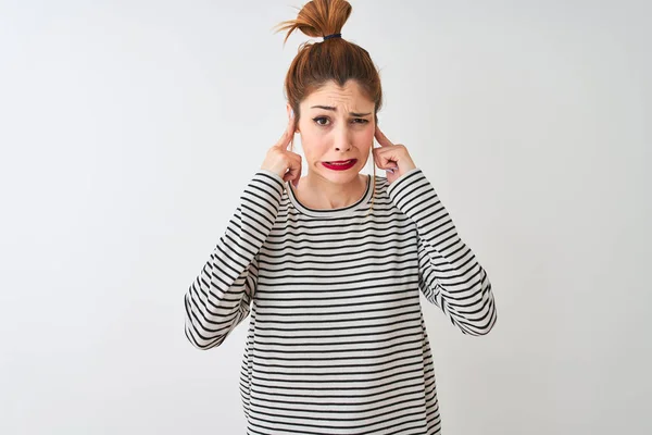 Redhead woman wearing navy striped t-shirt standing over isolated white background covering ears with fingers with annoyed expression for the noise of loud music. Deaf concept.