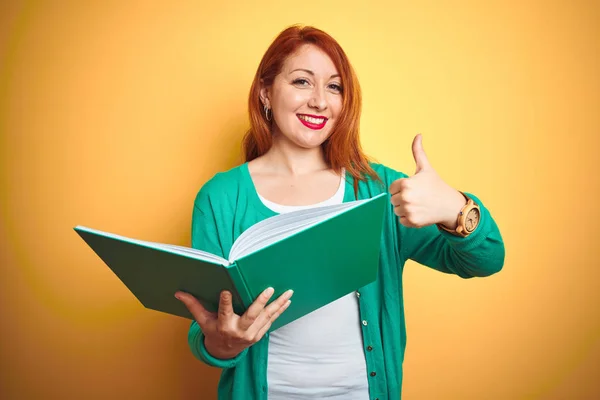 Young redhead student woman reading green book over yellow isolated background happy with big smile doing ok sign, thumb up with fingers, excellent sign
