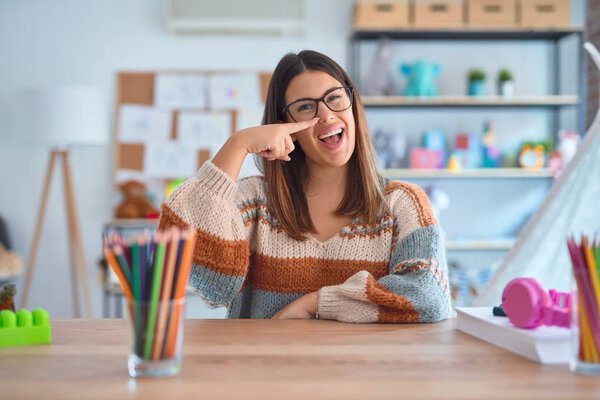 Young beautiful teacher woman wearing sweater and glasses sitting on desk at kindergarten Pointing with hand finger to face and nose, smiling cheerful. Beauty concept